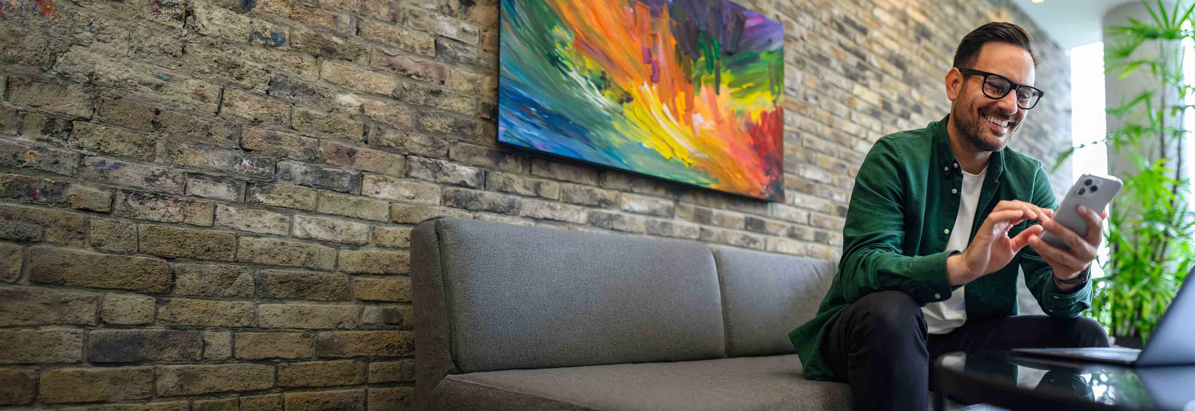 Man in a green shirt looking at his phone while sitting on a couch with a colorful oil painting on the wall behind him.
